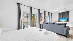 Ein Hotelzimmer mit Bett, grauem Sofa, Fernseher und Balkon mit Blick auf Palmen.