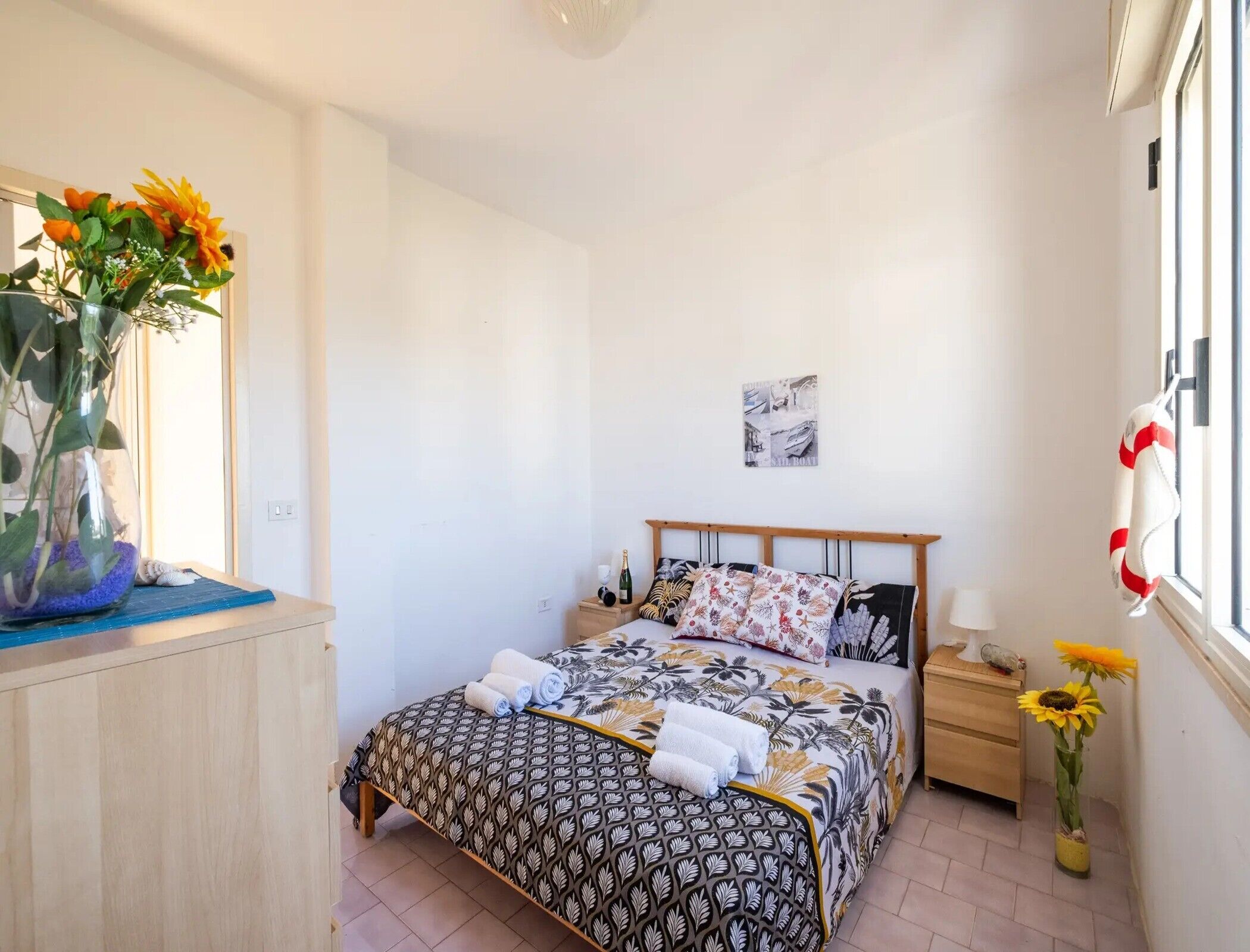 A bedroom with a bed, a nightstand, a vase with flowers, and a window with a towel hanging on the side.