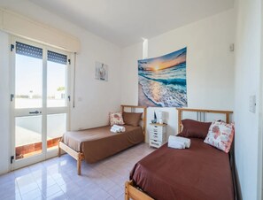 Ein Zimmer mit zwei Betten, einem Fenster mit Jalousien, einem Sonnenuntergangsgemälde und einem kleinen, weißen Nachttisch mit Lampe.