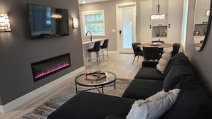 En moderne stue med en mørk sofa, et fladskærms-tv på væggen, en pejs og et spiseområde med bord og stole.