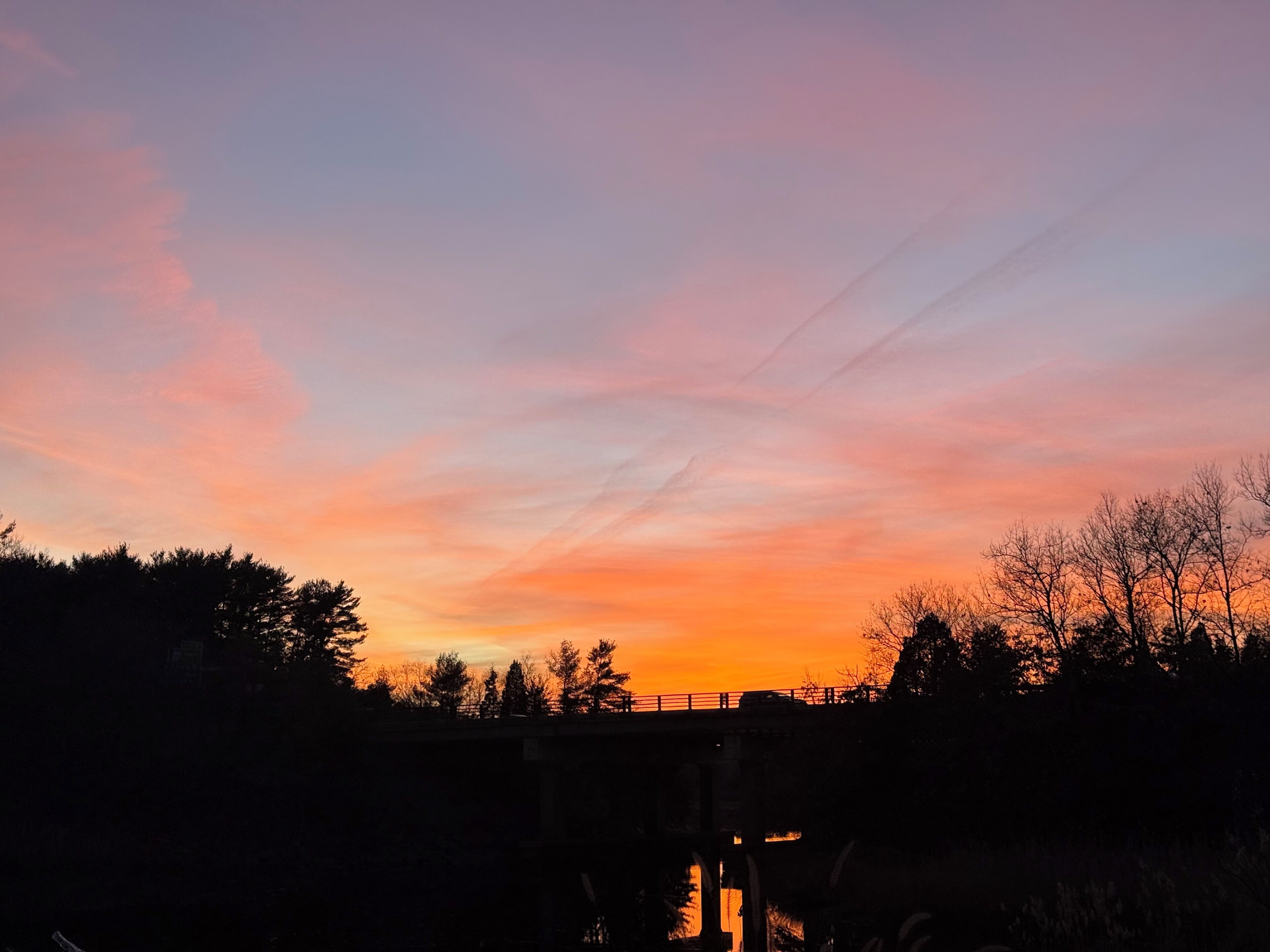 Een zonsondergang boven een brug, met silhouetten van bomen en een heldere lucht.