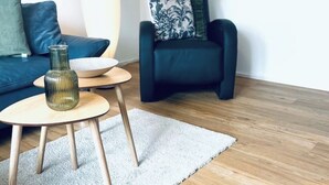 A modern living room with a blue sofa, a dark armchair, a small wooden table with a vase, and a white rug on a wooden floor.