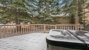 A wooden deck with a hot tub covered in snow, surrounded by trees and a building in the background. - I1 Gore Creek Meadows -Gorgeous Mountain Home, Private Hot Tub! (Vail)