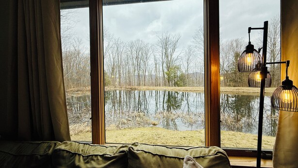 A room with a large window overlooking a body of water and trees, a wooden floor lamp, and a green sofa with cushions. - Stunning Pet Friendly House with Pond View, Sauna and Jetted Tub in the Home (Killington)