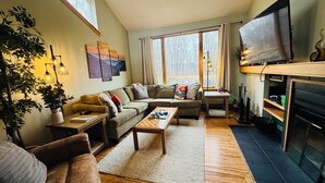 A cozy living room with a sofa, coffee table, and TV. There are plants, artwork, and a fireplace. - Stunning Pet Friendly House with Pond View, Sauna and Jetted Tub in the Home (Killington)