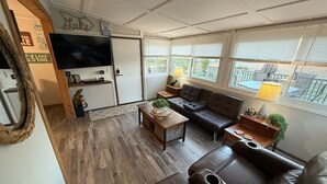 Un salon confortable avec un canapé, une table basse et un téléviseur. Il y a des fenêtres avec des stores, un plancher en bois et une porte avec une pancarte.