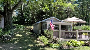 Une petite cabane avec une terrasse attenante, entourée d’une végétation luxuriante et d’arbres.