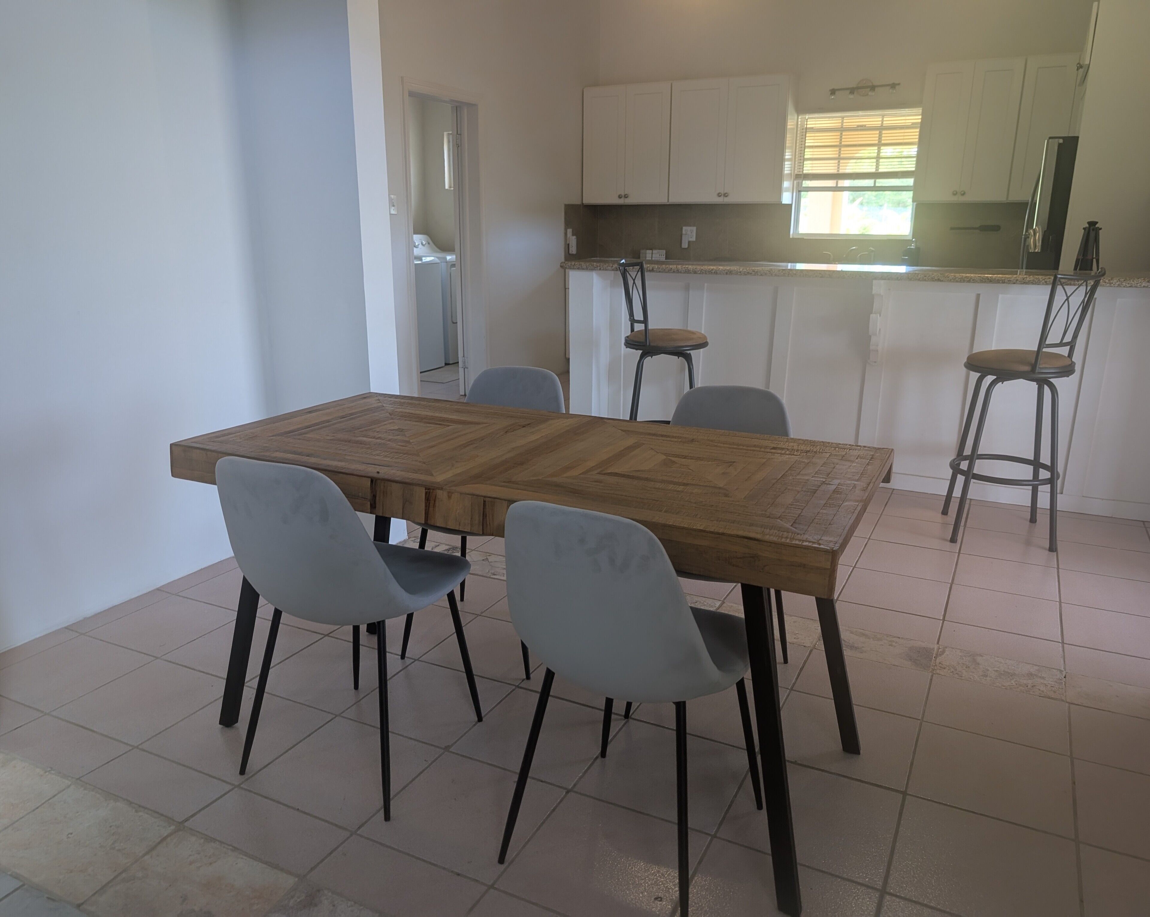 Un coin repas avec une table en bois et quatre chaises grises, une cuisine avec des meubles blancs et une fenêtre équipée de stores.