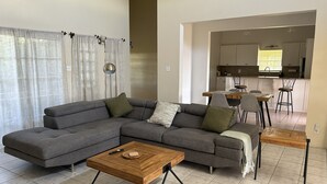 Een moderne woonkamer met een grijze hoekbank, een houten salontafel en een eethoek met een houten tafel en stoelen.