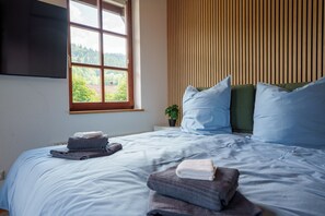 Un dormitorio con cama, ventana con vista a árboles, televisor montado en la pared y una planta en maceta.