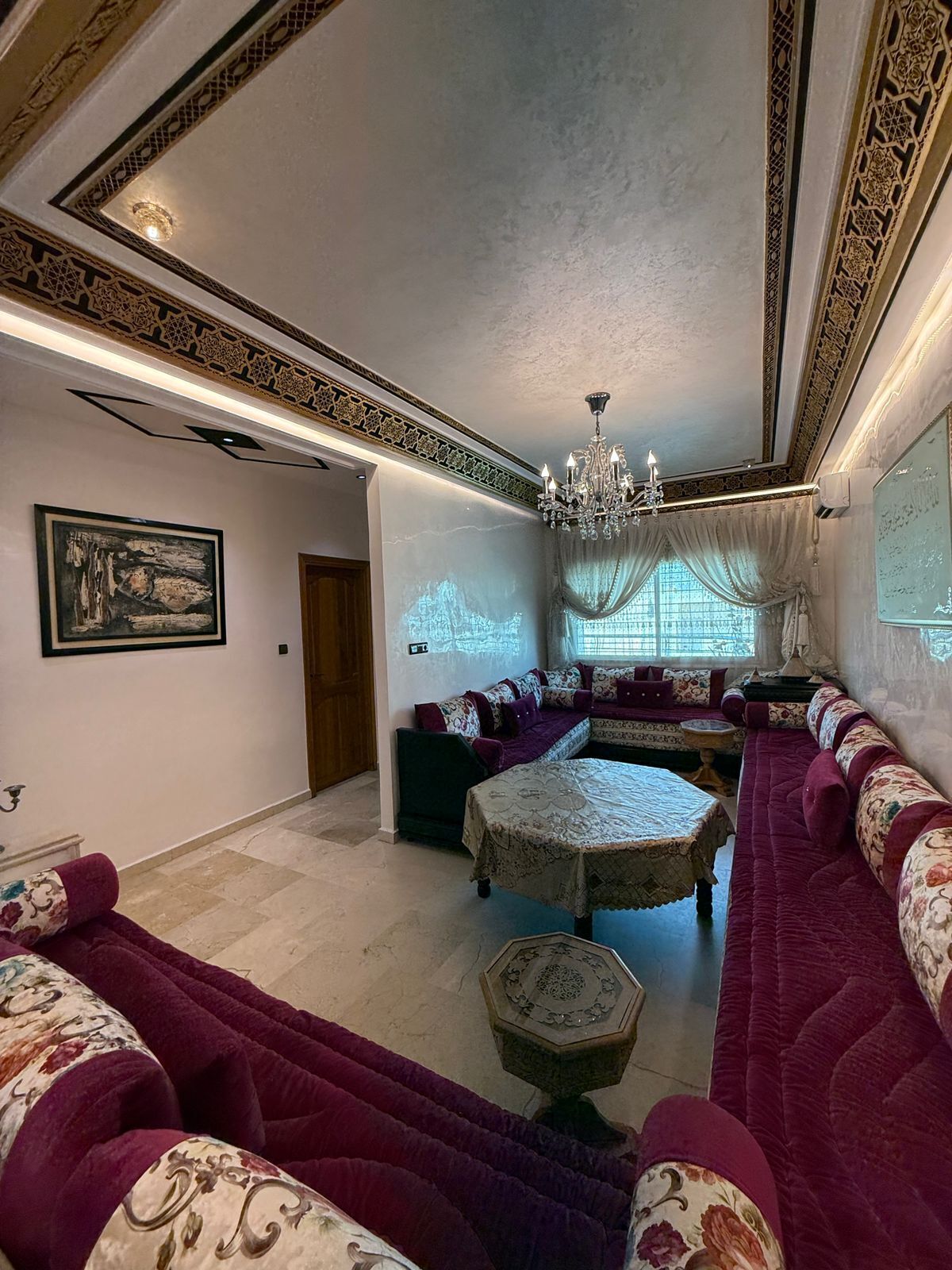 A room with a long, plush seating area, a chandelier, and patterned walls.