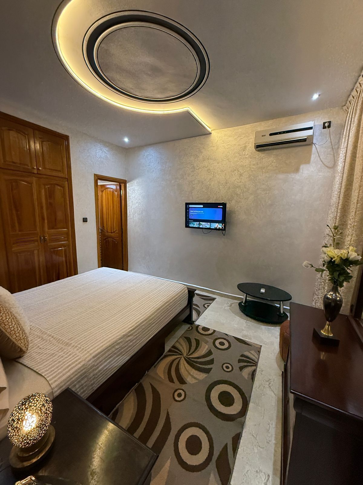Een moderne slaapkamer met een bed, een aan de muur bevestigde televisie en een klein tafeltje met een vaas.