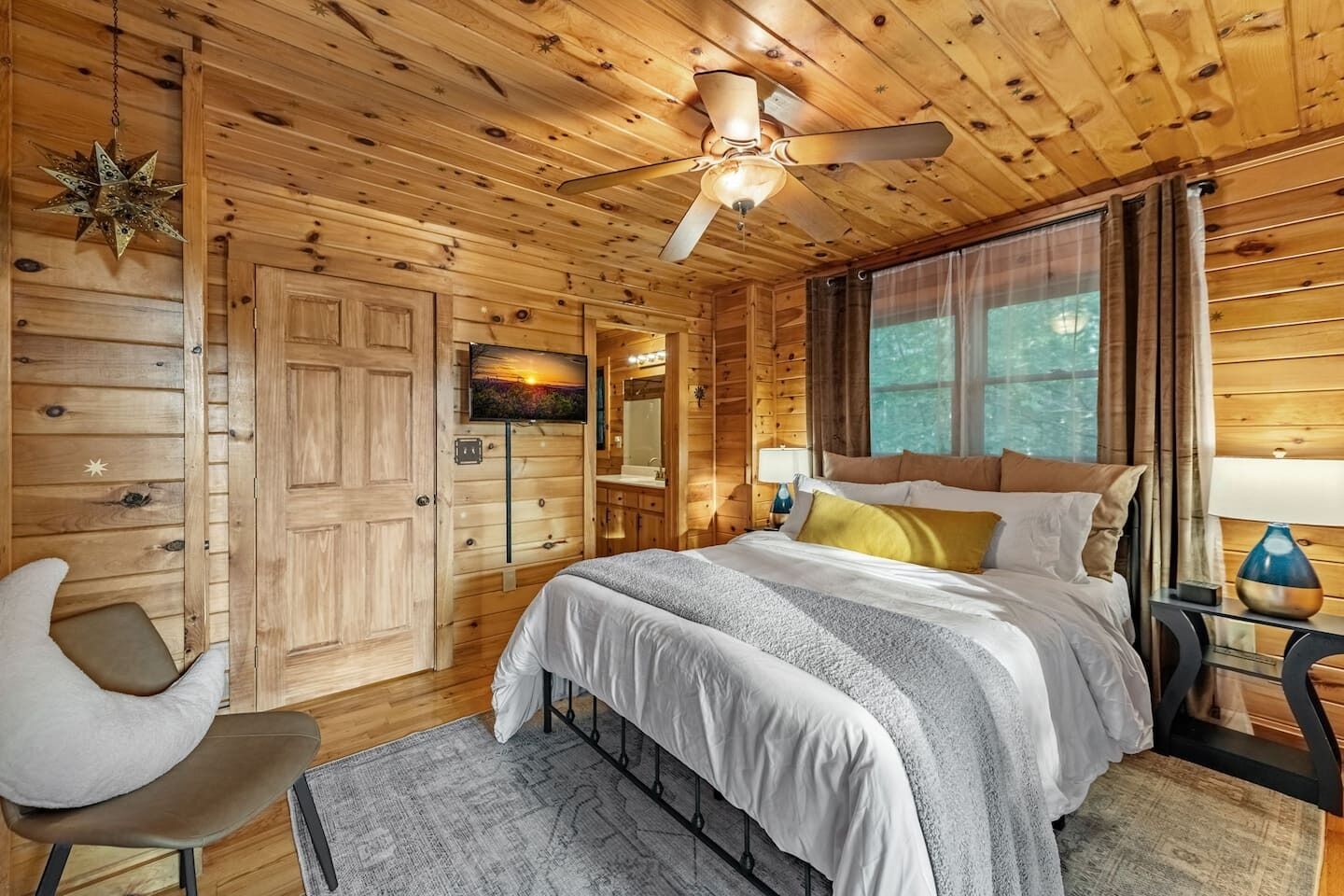 A wooden cabin bedroom with a bed, a chair, a ceiling fan, a TV, and a window with curtains.