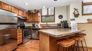 A kitchen with wooden cabinets, stainless steel appliances, a granite countertop, and a bar stool area. - Cozy 2 Bed Townhome with Fireplace + Noon Check In (Galena)