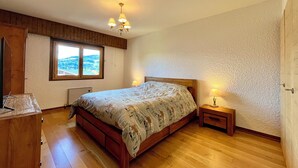 A bedroom with a wooden bed, a nightstand, a lamp, a window with a view, and a ceiling light. - The Balcony of the Giant in Combloux (Combloux)