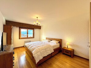 A bedroom with a wooden bed, a nightstand, a window with a view, and a ceiling light fixture. - The Balcony of the Giant in Combloux (Combloux)