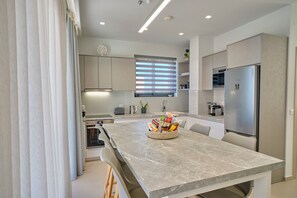 A modern kitchen with a marble countertop, built-in appliances, and a window with blinds. - Arillas Villas Dinos (Corfu)