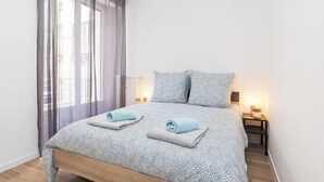 Una cama bien hecha con una colcha estampada, dos almohadas y toallas dobladas. Se ve una mesita de noche con una lámpara y un pequeño mueble auxiliar con otra lámpara.