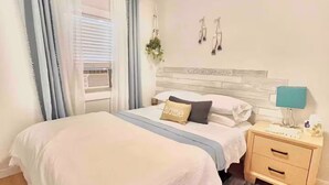 A bedroom with a large bed, a bedside table, a nightstand, a window with curtains, and a ceiling light fixture.