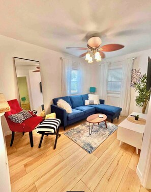 A modern living room with a blue sectional sofa, a red armchair, a striped chair, a small round table, and a ceiling fan. - Charming Coastal Retreat 1BR (Rockaway Beach)