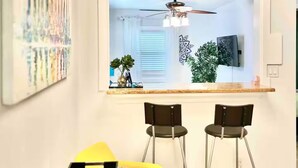 A modern kitchen with a dining area, featuring a ceiling fan, pendant lights, and a large window with blinds.