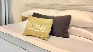 Un lit bien fait, avec un coussin décoratif à l’effigie d’une maison, une plante suspendue et un porte-serviettes fixé au mur.