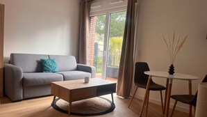 Una sala de estar moderna con un sofá gris, una mesa de centro de madera, una mesa redonda con sillas y vista a un edificio de ladrillo a través de grandes ventanales.