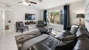 Een moderne woonkamer met een zwarte leren bank, een grijze fauteuil, een salontafel en een plafondventilator. Er is een groot raam met gordijnen, een flatscreen televisie aan de muur en een klein bijzettafeltje met een lamp.