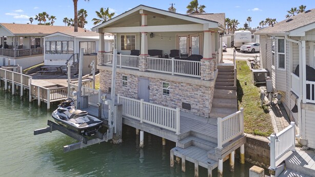 A waterfront property with a two-story house featuring a balcony, a dock with a boat, and a small boat house. - Stylish Canal-Front Home with Private Dock (Port Isabel)