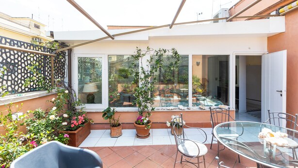 A sunlit patio with a glass table and chairs, surrounded by potted plants and a view of a garden. - Rental in Rome Rental in Rome Tre Lune Penthouse (Rome)