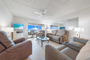A spacious living room with a large flat-screen TV, comfortable seating, a glass coffee table, and a ceiling fan. - Tivoli by the Sea (Siesta Key)