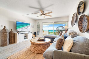 A spacious living room with a large flat-screen TV, a ceiling fan, wicker furniture, and a view of the ocean through large sliding doors. - Tivoli by the Sea (Siesta Key)