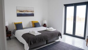 Ein modernes Schlafzimmer mit einem großen Bett, zwei Nachttischlampen, Blick durch eine Schiebetür und einem Gemälde an der Wand.