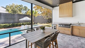A modern kitchen with a large island, a dining area with a pool view, and a barbecue grill. - Neptune of the Sea, sleeps 12 with a Pool (St Simons Island)