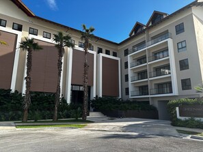 Un edificio moderno de varios pisos con una entrada central, rodeado de palmeras y follaje.
