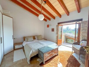Une chambre avec un plafond en bois, un lit recouvert d’une courtepointe blanche et bleue, une commode en bois, une porte blanche et un balcon agrémenté de meubles en osier.