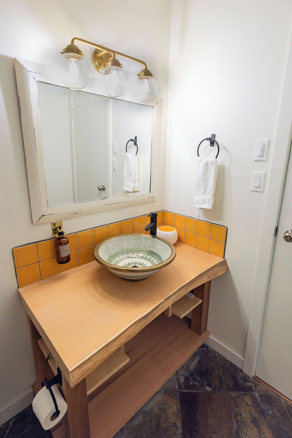 Een badkamer met een houten wastafelmeubel, een rond wastafeltje, een spiegel met gouden lijst en een handdoekrek.