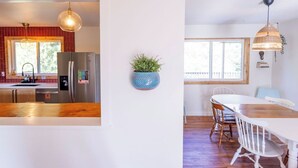 Una cocina moderna con refrigerador, pileta y encimera de madera. Hay un comedor con una mesa y sillas blancas, y una lámpara colgante.