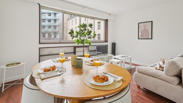 A modern living room with a round wooden dining table set for breakfast, a sofa, a TV, and a large window with a view of buildings. - Chic 1BR Steps from Opéra Garnier (Paris)