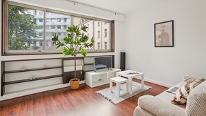 A modern living room with a large window, a flat-screen TV, a white sofa, and a small table. - Chic 1BR Steps from Opéra Garnier (Paris)