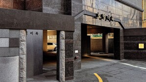 Una entrada de edificio con un letrero en coreano, un área de estacionamiento y una línea amarilla en el pavimento.