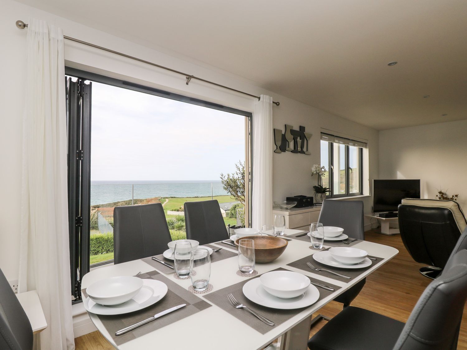 A dining area with a table set for four, large windows offering a view of the sea, and a TV on a stand to the right.