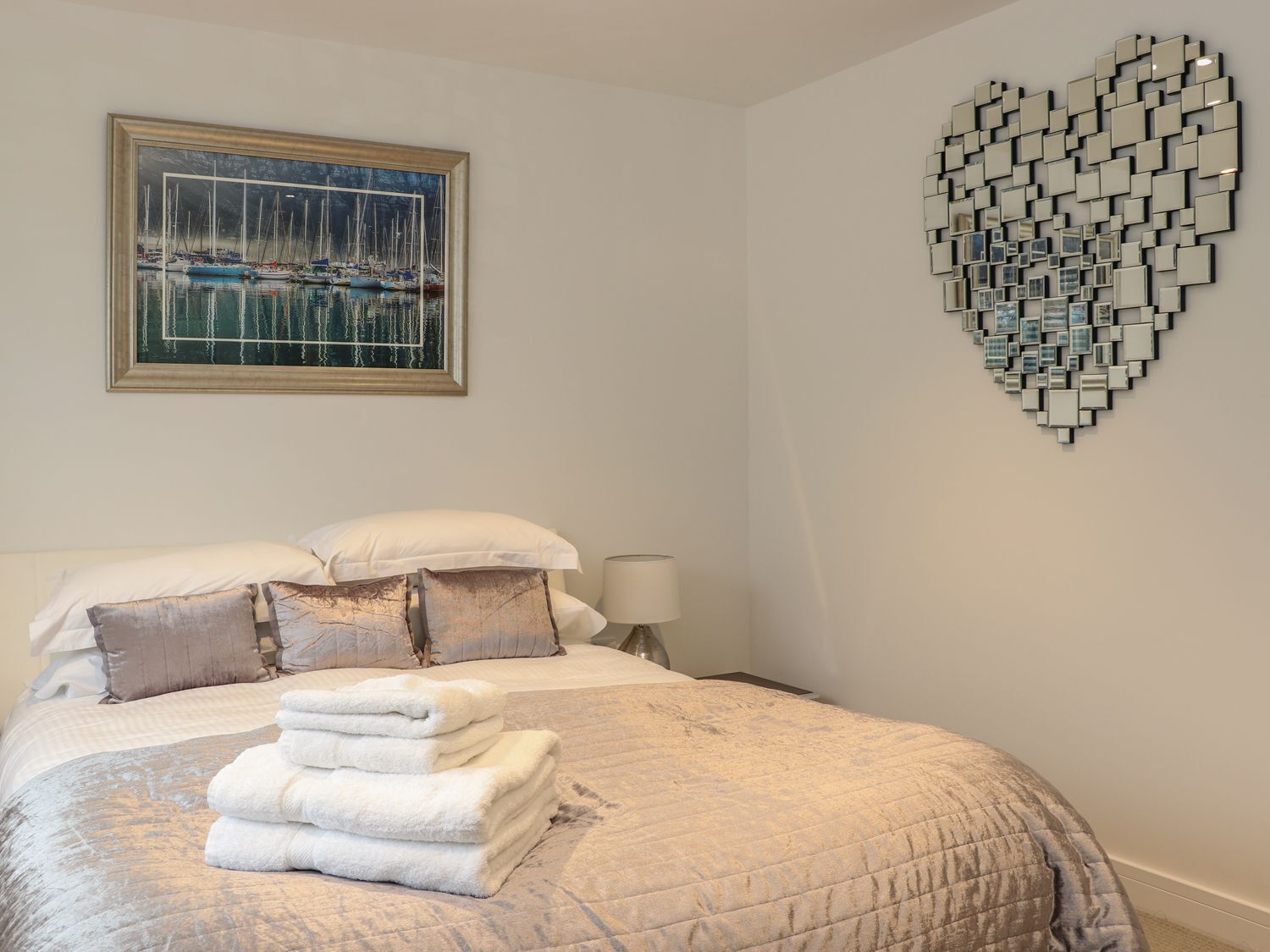 A neatly made bed with a quilted cover, a stack of white towels, and a painting of a marina above it.