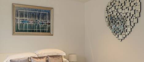 A neatly made bed with a quilted cover, a stack of white towels, and a painting of a marina above it.