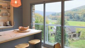 A modern kitchen with a large island, bar stools, and a view of a green landscape through large windows.
