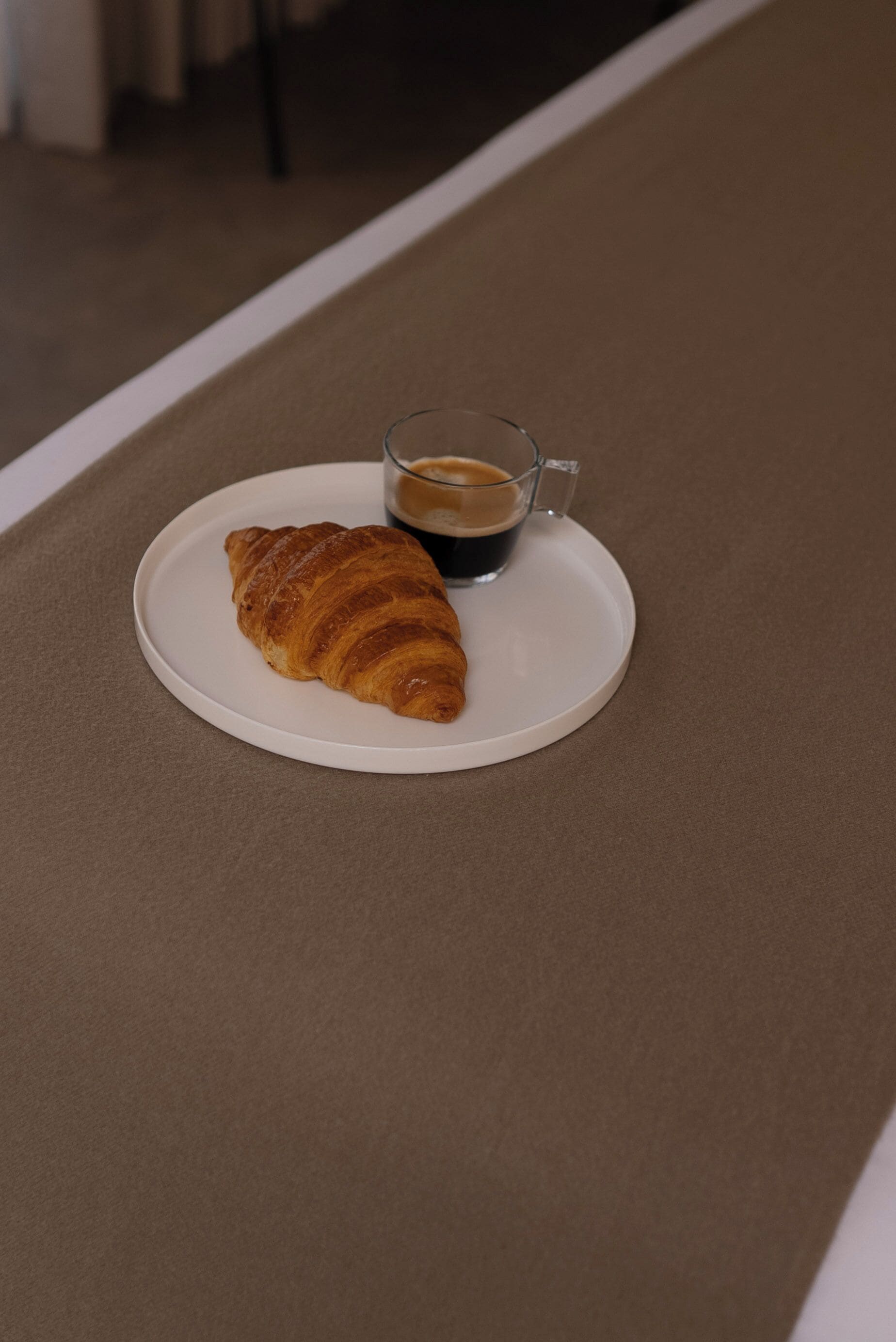 Un croissant sobre un plato blanco, junto a una taza de café, todo sobre una mesa marrón.