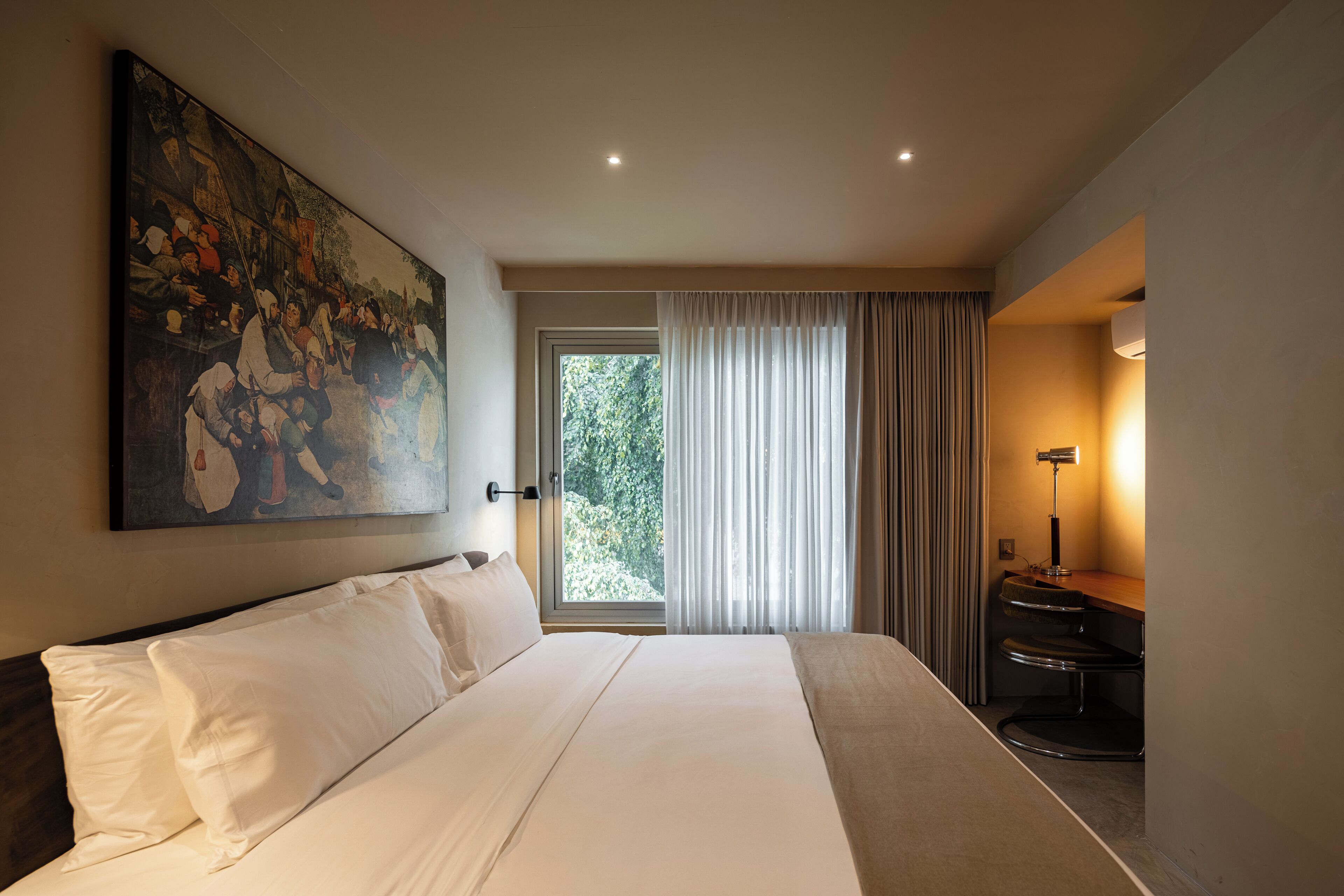A hotel room with a large bed, a painting on the wall, a window with curtains, and a desk with a lamp.