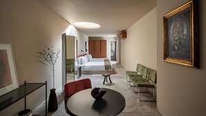A modern hotel room with a bed, a dining area with a table and chairs, a sofa, a painting on the wall, and a vase with branches.