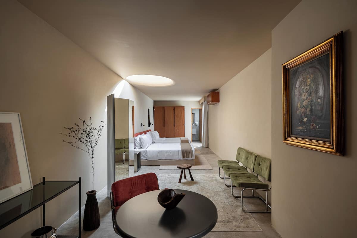 Une chambre d’hôtel moderne comprenant un lit, un coin repas avec une table et des chaises, un canapé, un tableau au mur et un vase avec des branches.