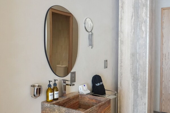 A bathroom with a large oval mirror, a wooden vanity, two bottles of hair products, a hair dryer, and a small metal cup.